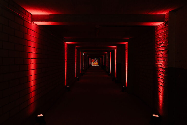 Ein dunkler Flur, der von roten Lichtern entlang der Backsteinwände beleuchtet wird und so einen dramatischen, tunnelartigen Effekt erzeugt - perfekt für Kultur & private Feiern bei Catering im BASE Köln - Eventlocation für Business.