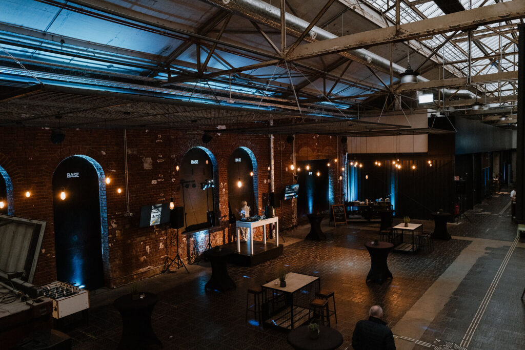Veranstaltungsraum im Industriestil mit freigelegten Backsteinwänden, Lichterketten und im Raum verteilten Tischen - ideal für kulturelle & private Feiern oder Catering im BASE Köln - Eventlocation für Business.