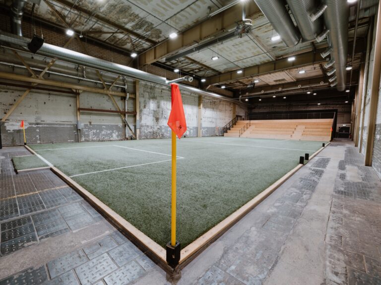 Ein Hallenfußballfeld mit Kunstrasen, orangefarbenen Eckfahnen und leeren Tribünen im Hintergrund.