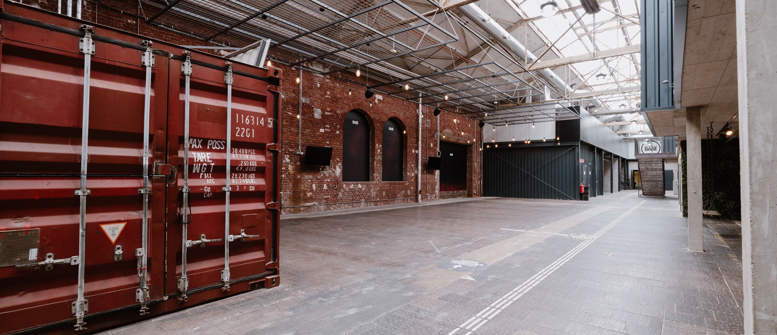 Ein geräumiger, industriell anmutender Flur mit Backsteinwänden, Oberlichtern und einem roten Frachtcontainer auf der linken Seite.