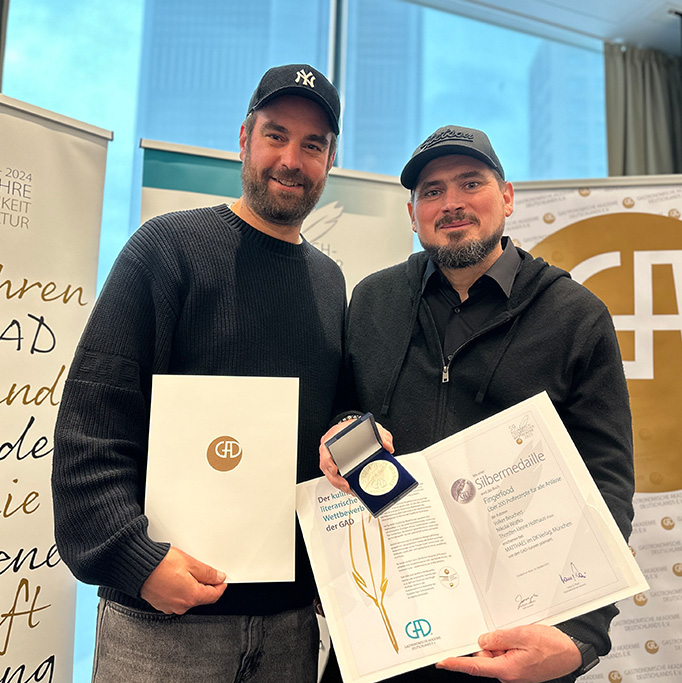 Zwei Männer lächeln, halten eine Urkunde und eine Medaille für Nachhaltigkeit im Catering in der Hand und stehen vor einem Preisbanner.
